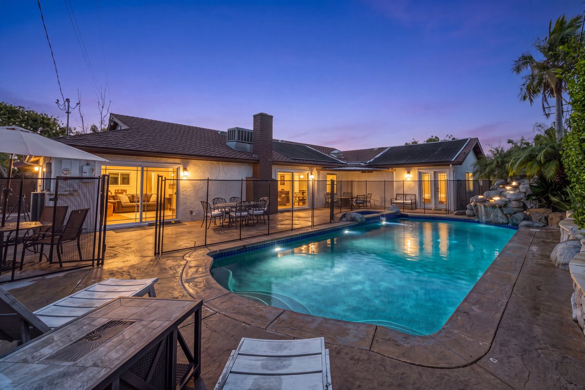 The backyard at dusk: the swimming pool and hot tub behind a safety fence, with a patio and a rock waterfall